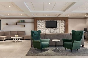 A modern living room with a sectional sofa, two green armchairs, a small round table, and a flat-screen TV mounted on a geometric-patterned wall. - Wyndham Garden Totowa (Totowa)