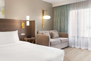 A hotel room with a bed, a sofa, a small table, and a wall-mounted lamp.