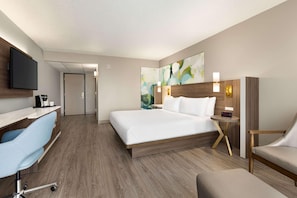 A hotel room with a bed, desk, chair, TV, and artwork on the wall.