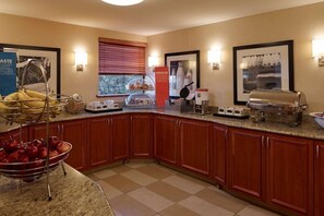 Uma cozinha de hotel com bancada de granito, armários de madeira, uma tigela de frutas, um espaço para café e um quadro com a imagem de uma máquina de café.