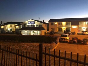 Un motel con un llamativo letrero que dice 'MOTEL', varios espacios de estacionamiento y un área cercada al frente.