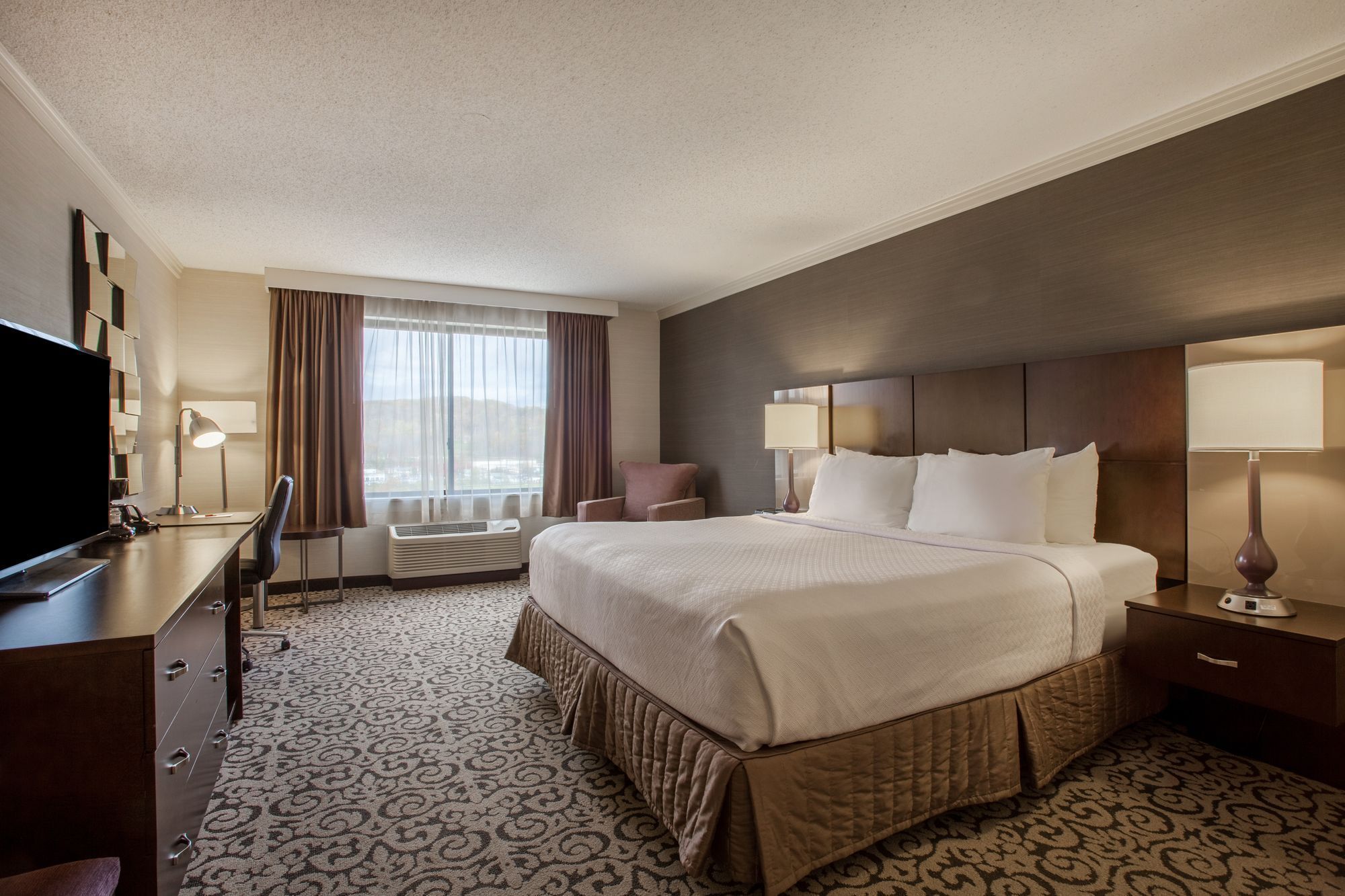 A hotel room with a large bed, a desk with a TV, a chair, a window with curtains, and a wall-mounted lamp.