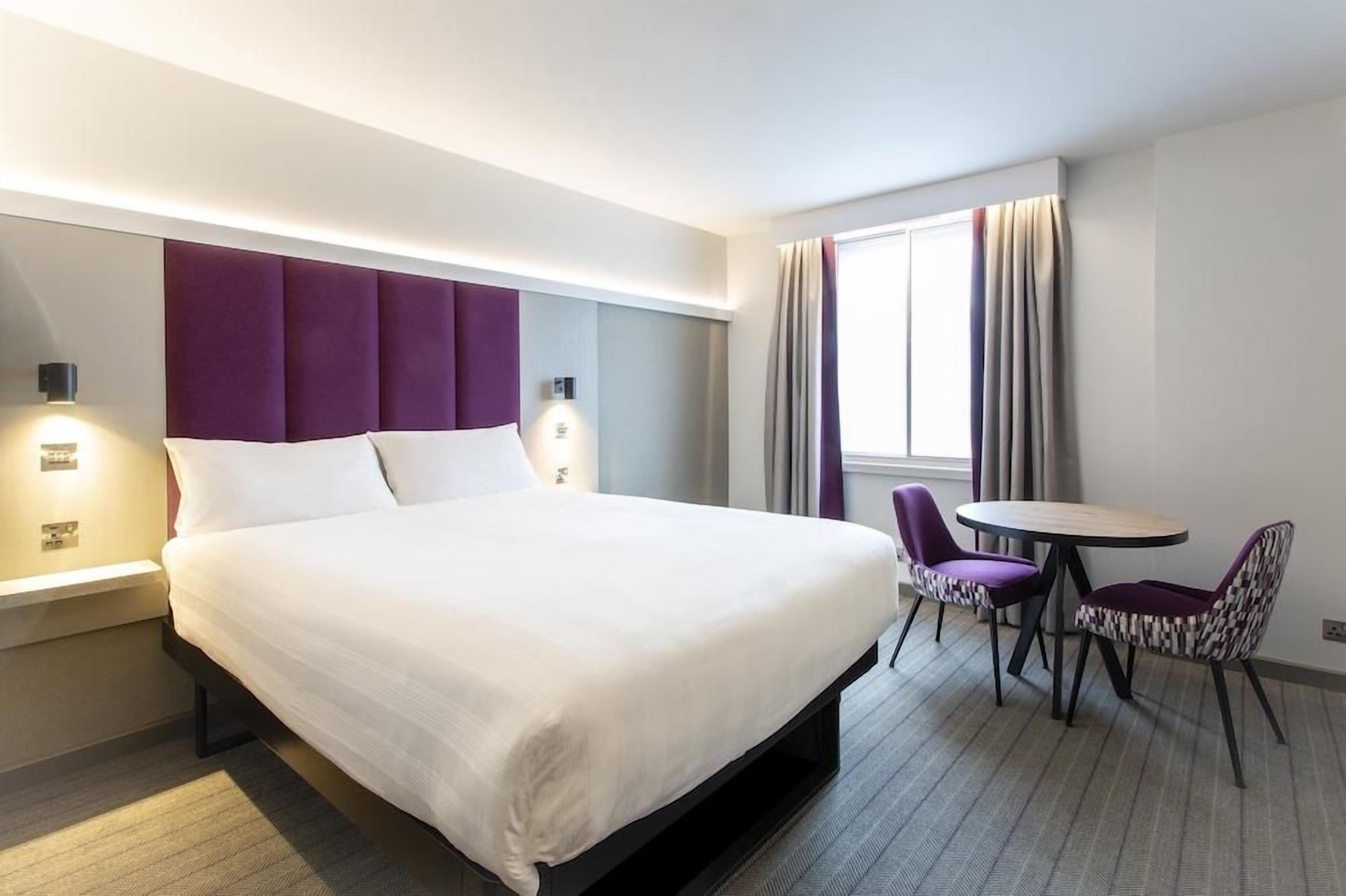 A hotel room with a large bed, a bedside table, a chair, a small table, and a window with curtains.