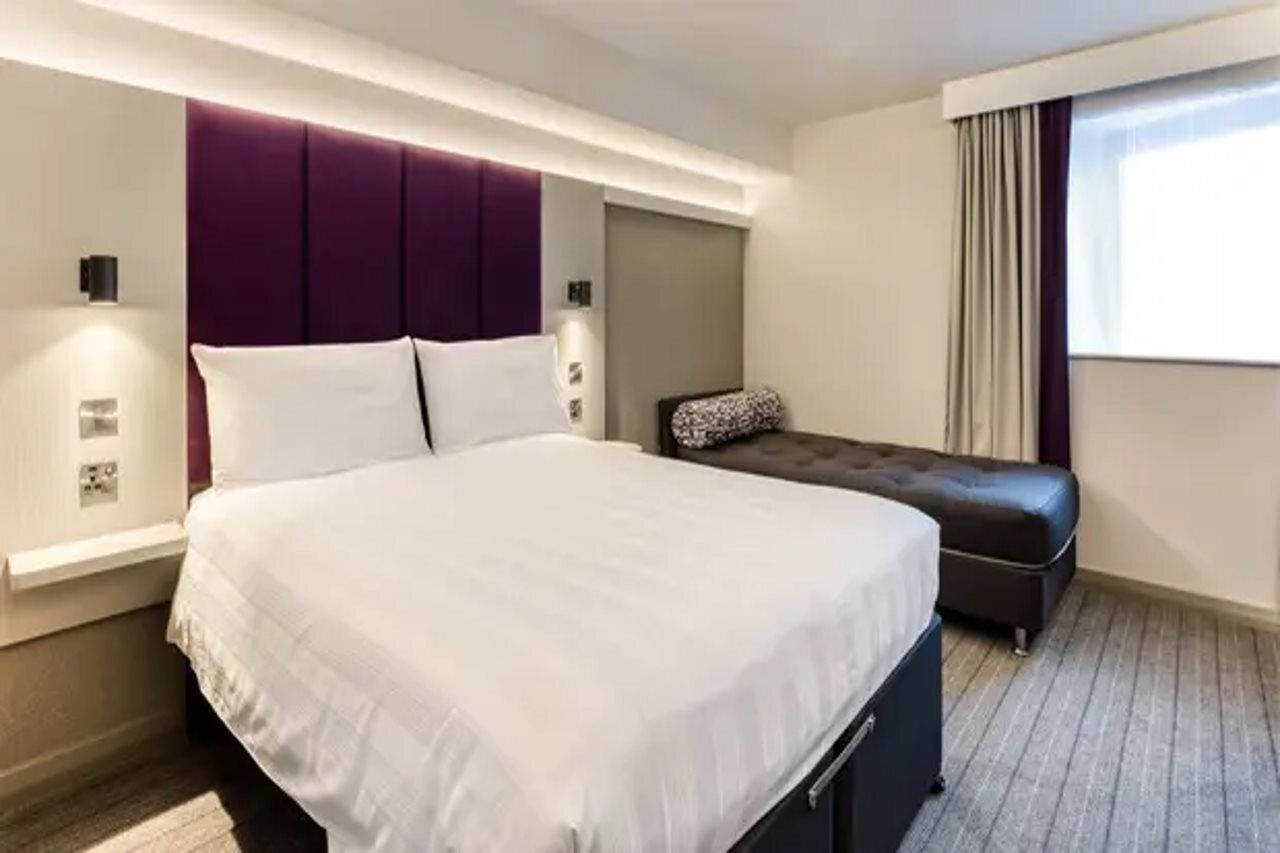 A hotel room with a large bed, a padded bench, a wall-mounted light, and a window with curtains.