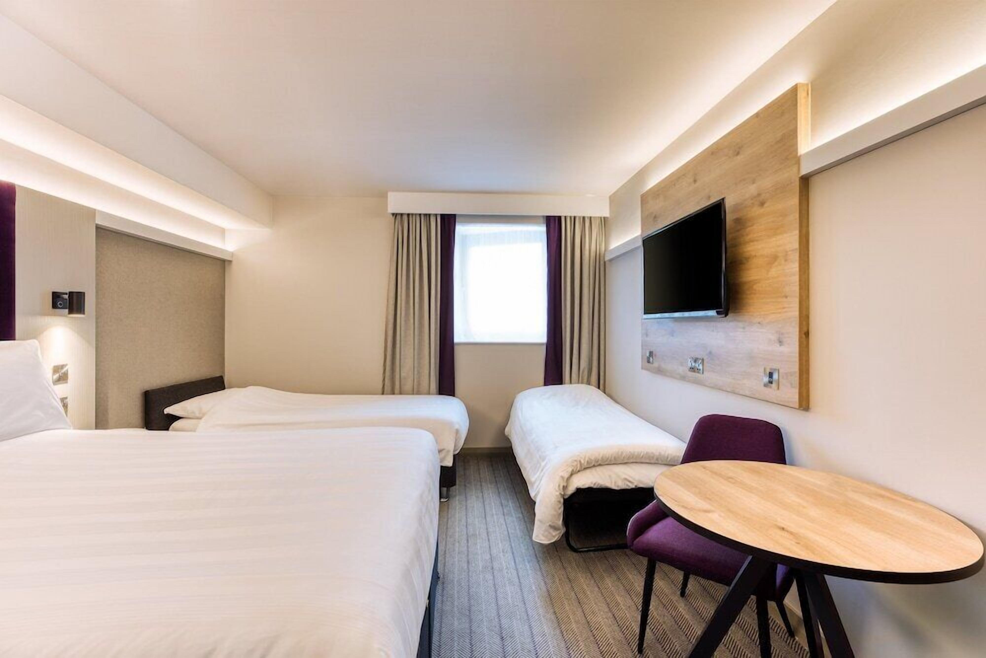 A hotel room with two single beds, a small table, a TV mounted on the wall, and a window with curtains.