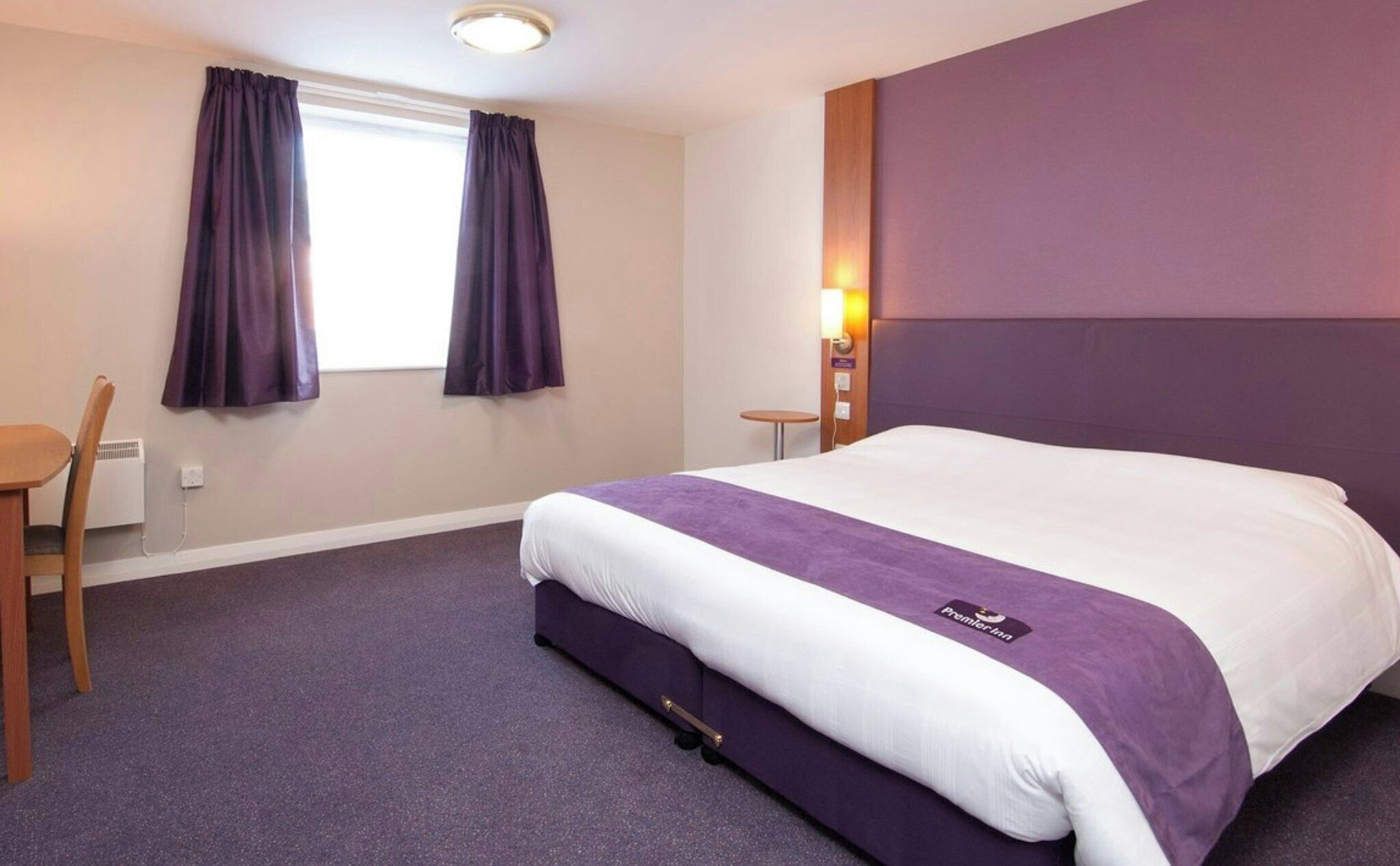 A hotel room with a bed, a chair, a small table, and a window with curtains.