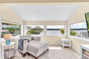 A living room with a sofa, a small table with a lamp, a side table, and a bookshelf. There are three large windows offering views of a body of water and greenery.