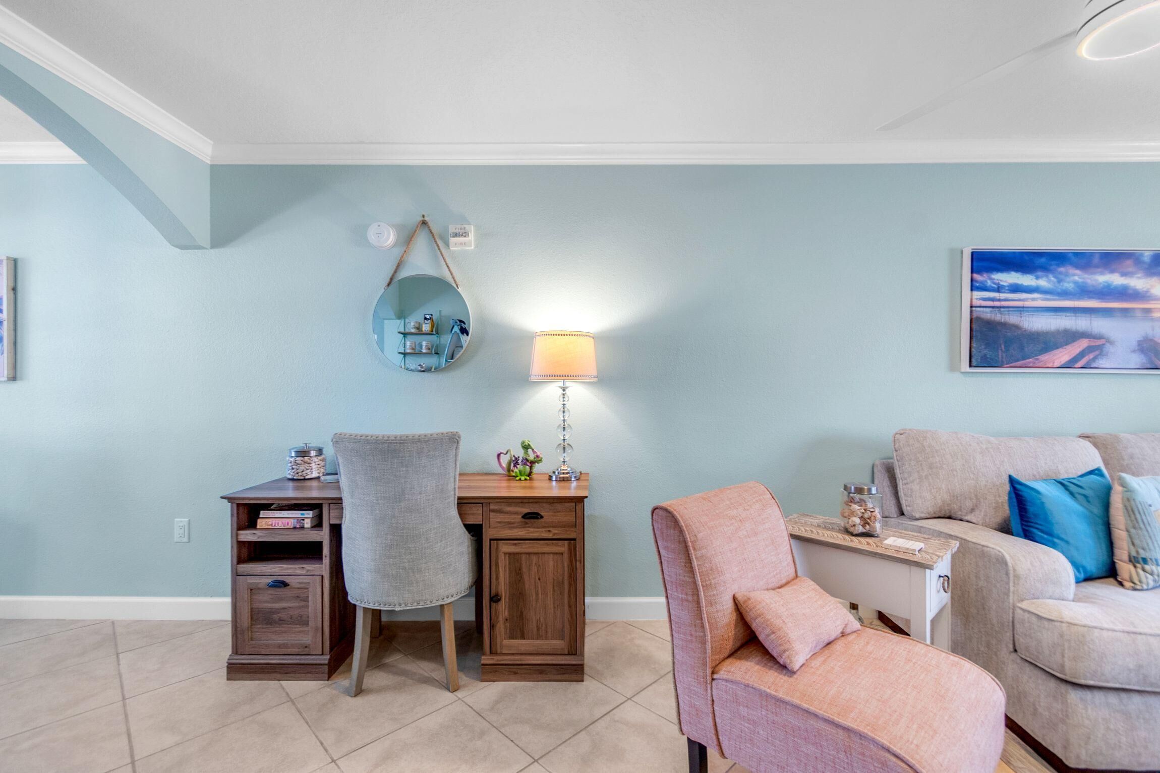 A room with a desk, chair, and couch, a lamp, a round mirror, and a painting on the wall.