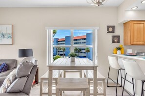 A modern kitchen with a white table and bar stools, a sofa with a throw blanket, and a window with a view of buildings and palm trees. - Skyline of Madeira 306 (Madeira Beach)