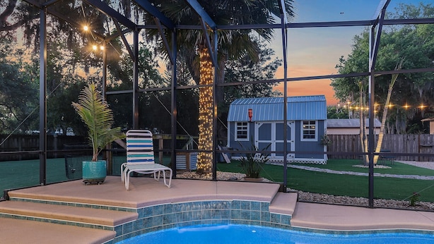 A swimming pool with steps leading up to it, a covered patio area with a chair and a potted plant, and a small house with string lights in the background. - Sunny Dayz (New Smyrna Beach)