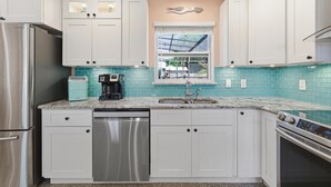A kitchen with white cabinets, a stainless steel refrigerator, a dishwasher, a coffee maker, and a window with a view of the outside. - Sunny Dayz (New Smyrna Beach)