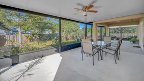 Eine geräumige Terrasse mit Esstisch und Stühlen, einem Deckenventilator und großen Fenstern mit Blick auf einen Garten.