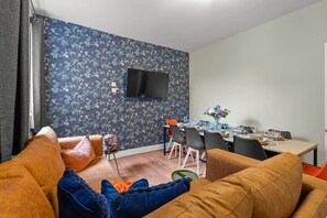 Una sala de estar con un sofá, un comedor con una mesa dispuesta para cuatro personas, un televisor de pantalla plana en la pared y un papel tapiz estampado.