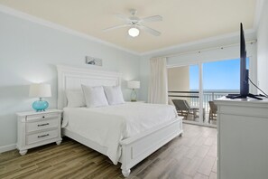 Quarto com cama branca, mesa de cabeceira com abajur, ventilador de teto e varanda com cadeiras.