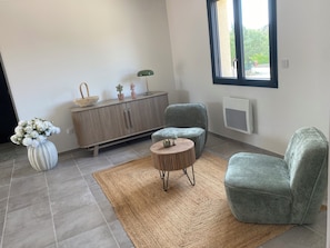 A room with a grey sofa, a small round table, a cabinet with decorative items, and a window with a view of greenery. - House Swap Orgnac l'aven (Orgnac-l'Aven)