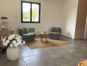 A modern living room with two grey armchairs, a round coffee table, a vase with white flowers, a wooden side table, and a large window with a view of trees.