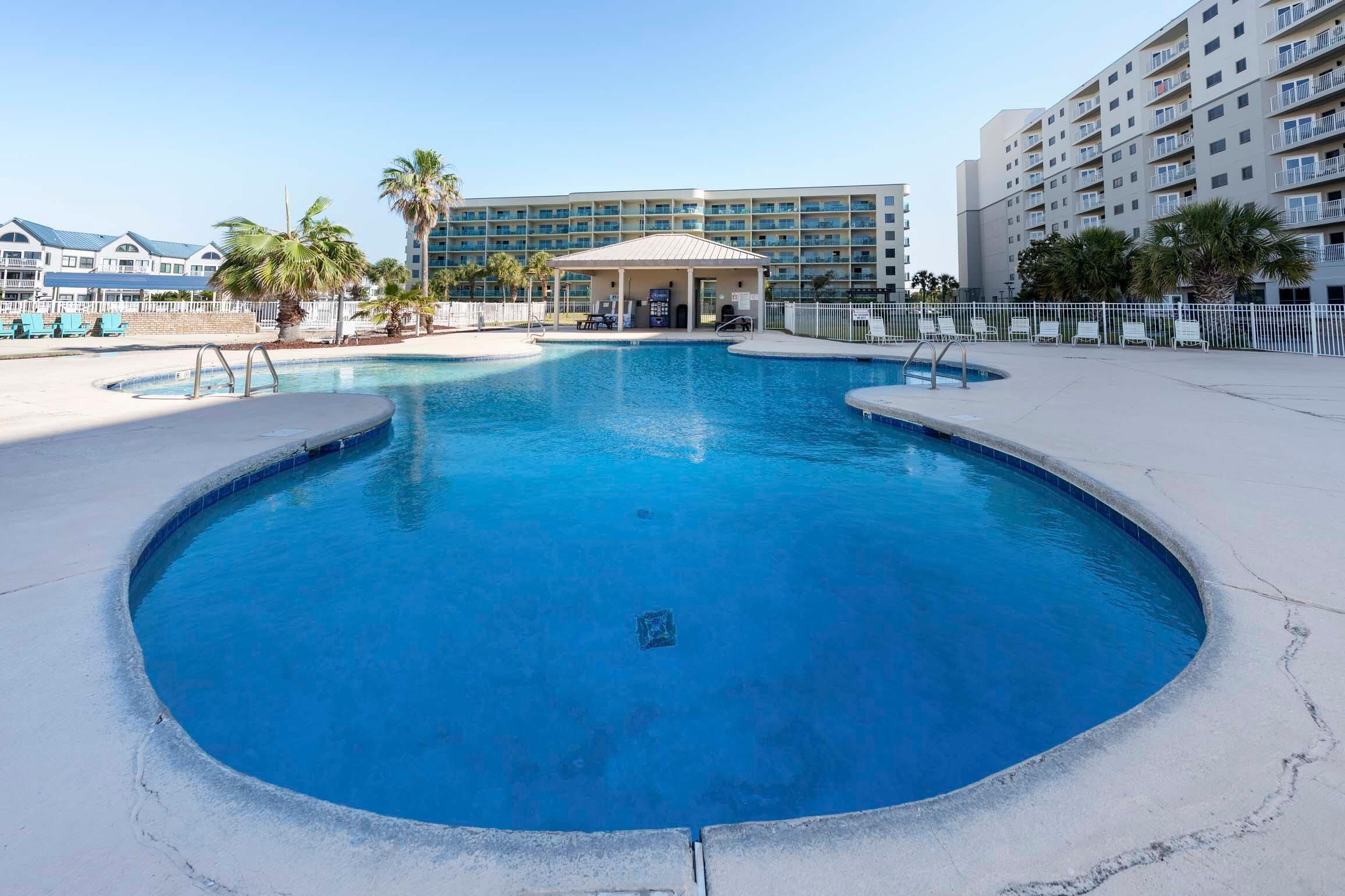 A swimming pool with a covered area, surrounded by a fence, and adjacent to a multi-story building with balconies.