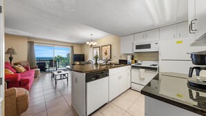 Een compacte keuken met witte kasten, een vaatwasser en een fornuis. Er is een zithoek met een rode bank en een kleine tafel. Een balkon met uitzicht op de oceaan is zichtbaar.