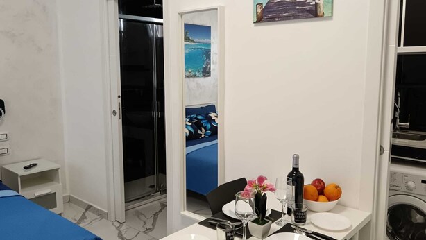 A hotel room with a bed, a dining table set with plates, glasses, and a bottle, a washing machine, and a painting of a pier. - The Colors of the Sea - Turquoise Studio Apartment (San Pietro In Bevagna)