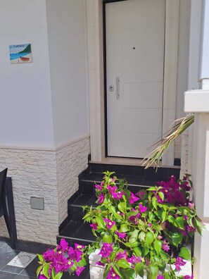 Eine weiße Tür mit einem kleinen Schild darüber, eine Treppe aus schwarzen Steinen, die zur Tür hinaufführt, und ein Blumentopf mit violetten Blüten im Vordergrund.