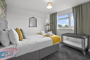 Habitación de hotel con cama, cuna, ventana con cortinas, espejo colgado y lámpara de noche.
