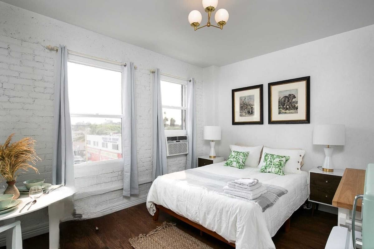 A bedroom with a bed, two bedside tables with lamps, a window with curtains, and two framed pictures on the wall.