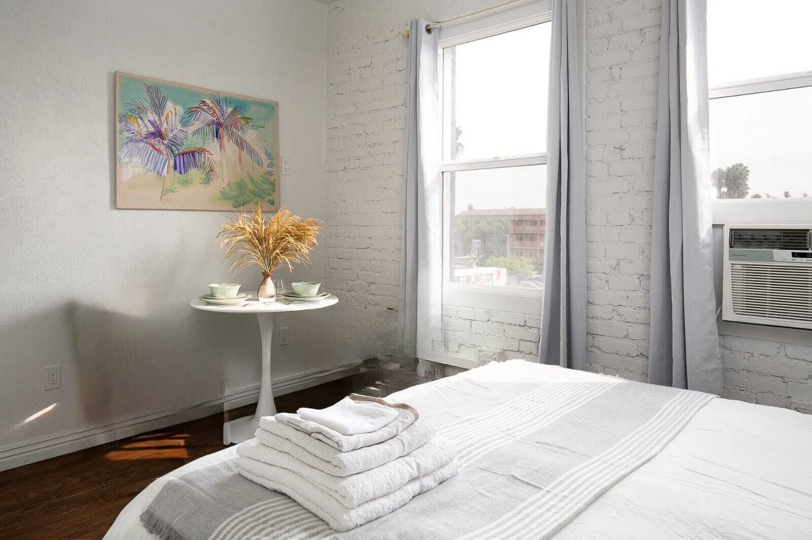 A hotel room with a bed, a small table with a vase and cups, a painting on the wall, and a window with curtains.