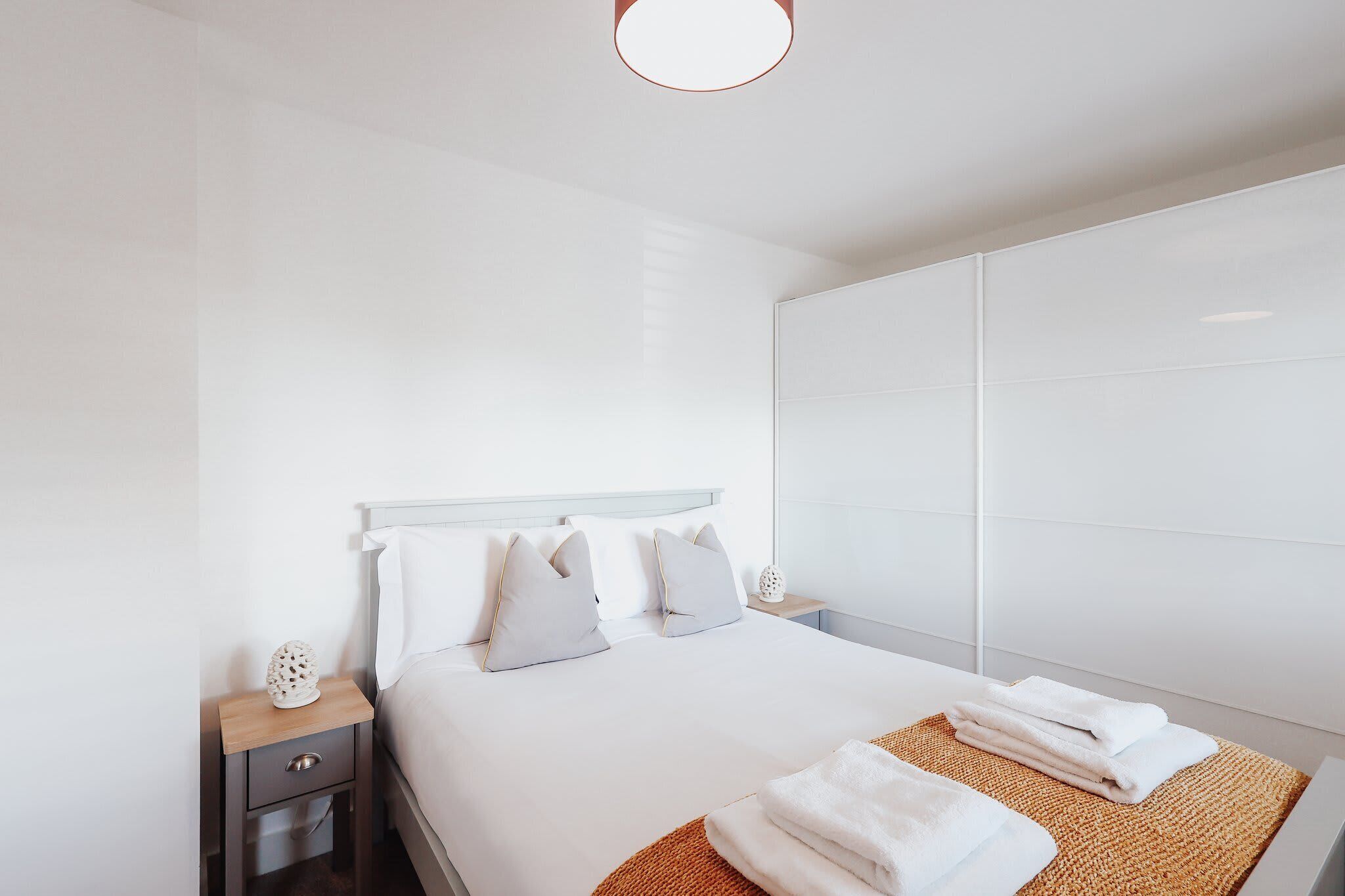 A modern hotel room with a large bed, a bedside table with a lamp, and a large mirror.