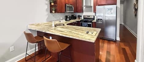 A kitchen with a marble countertop, stainless steel appliances, wooden cabinets, and two bar stools.