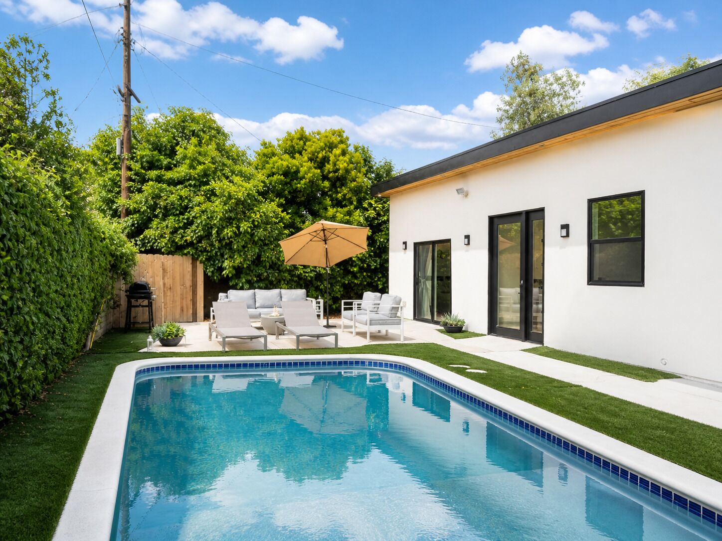 Une piscine avec un bord carrelé, un bâtiment blanc aux fenêtres à encadrement noir et une clôture en bois.