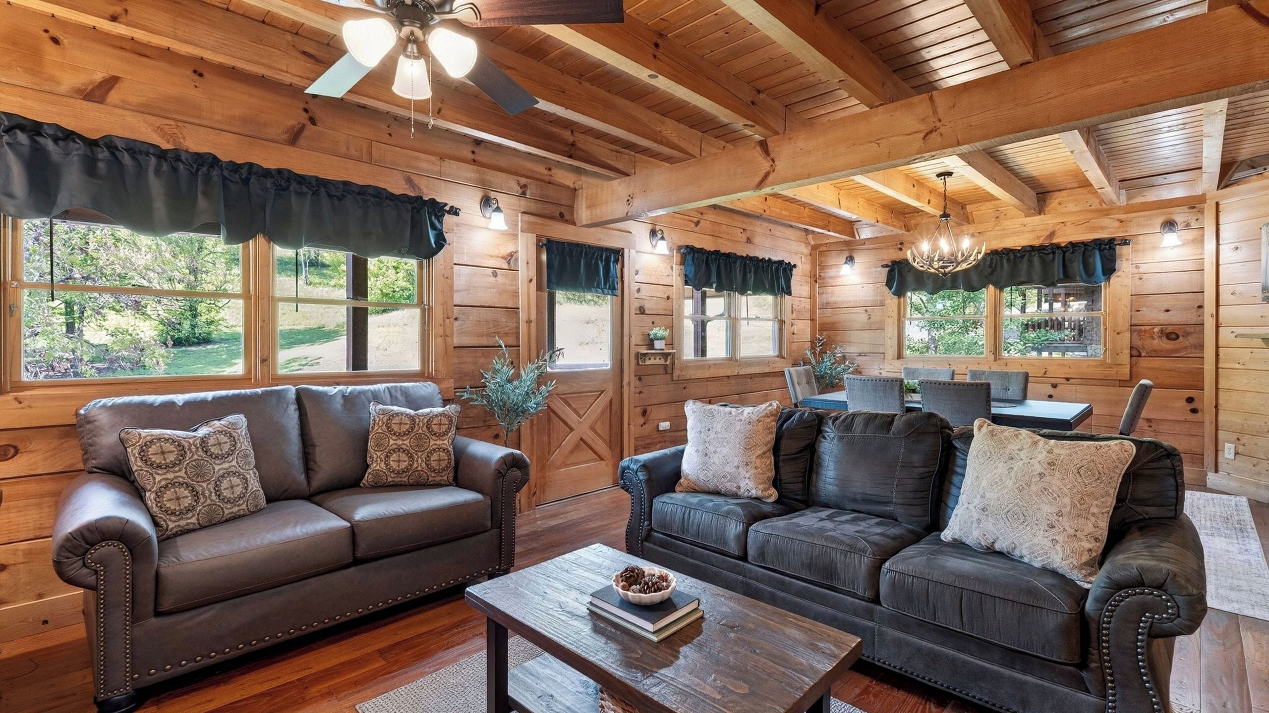 A log cabin interior with two leather sofas, a wooden coffee table, and a dining area with a chandelier.