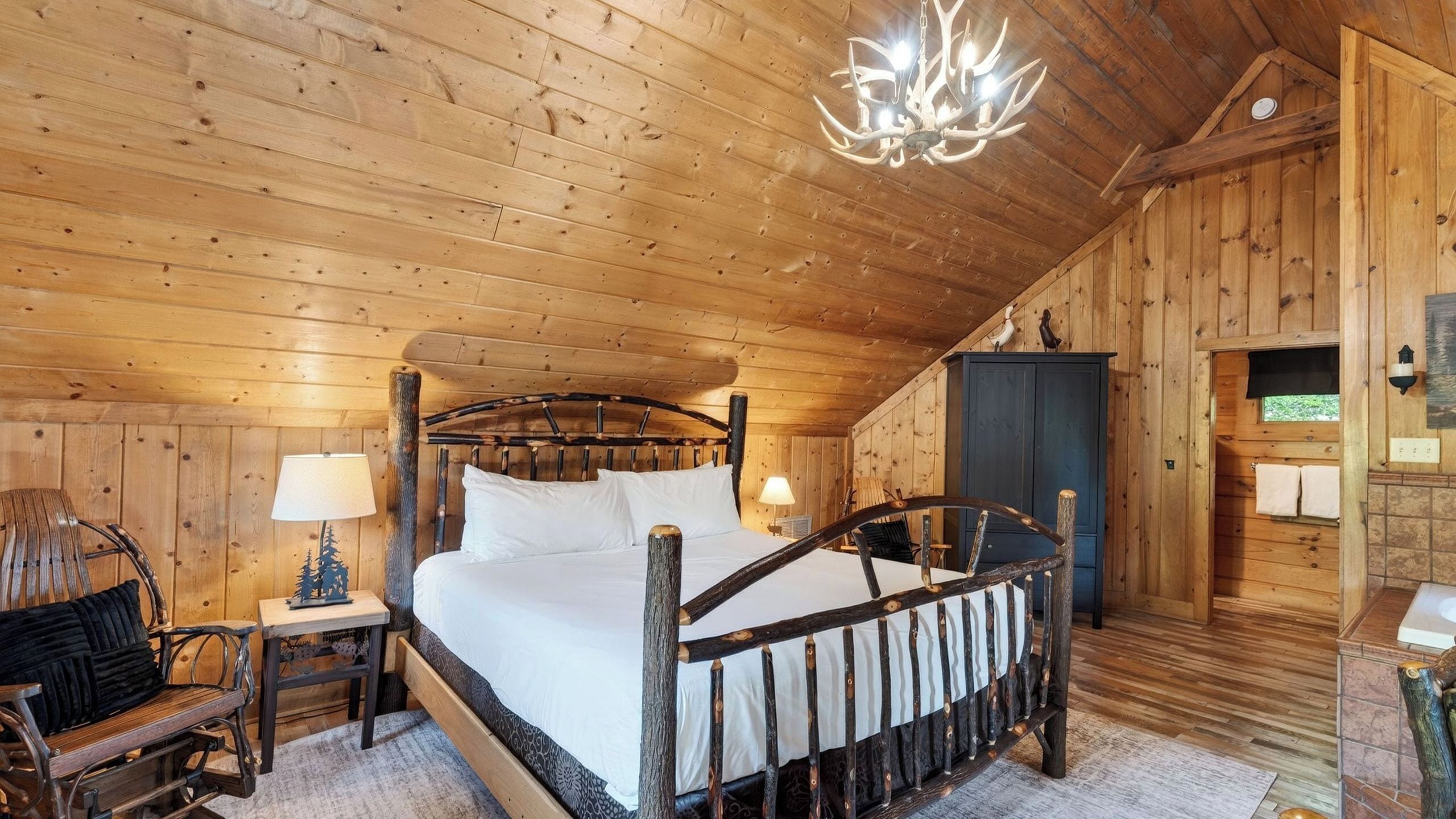 A wooden cabin bedroom with a bed, nightstands, a chair, a chandelier, and a wardrobe.