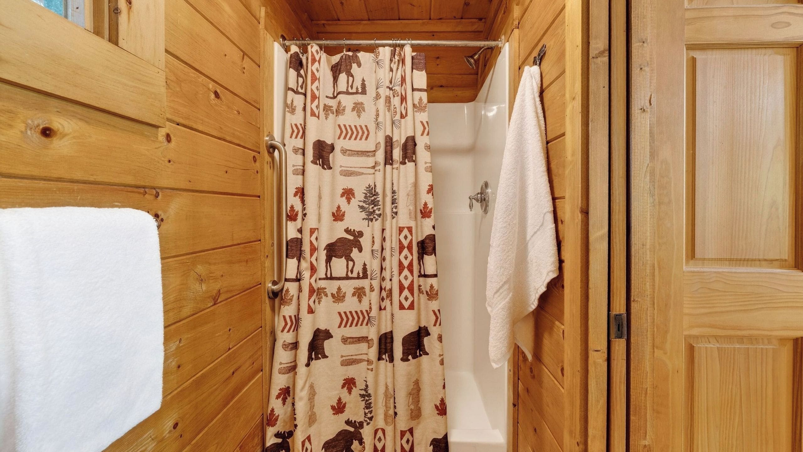 A wooden cabin bathroom with a shower curtain featuring animal prints, a white towel hanging on the wall, and a wooden door.