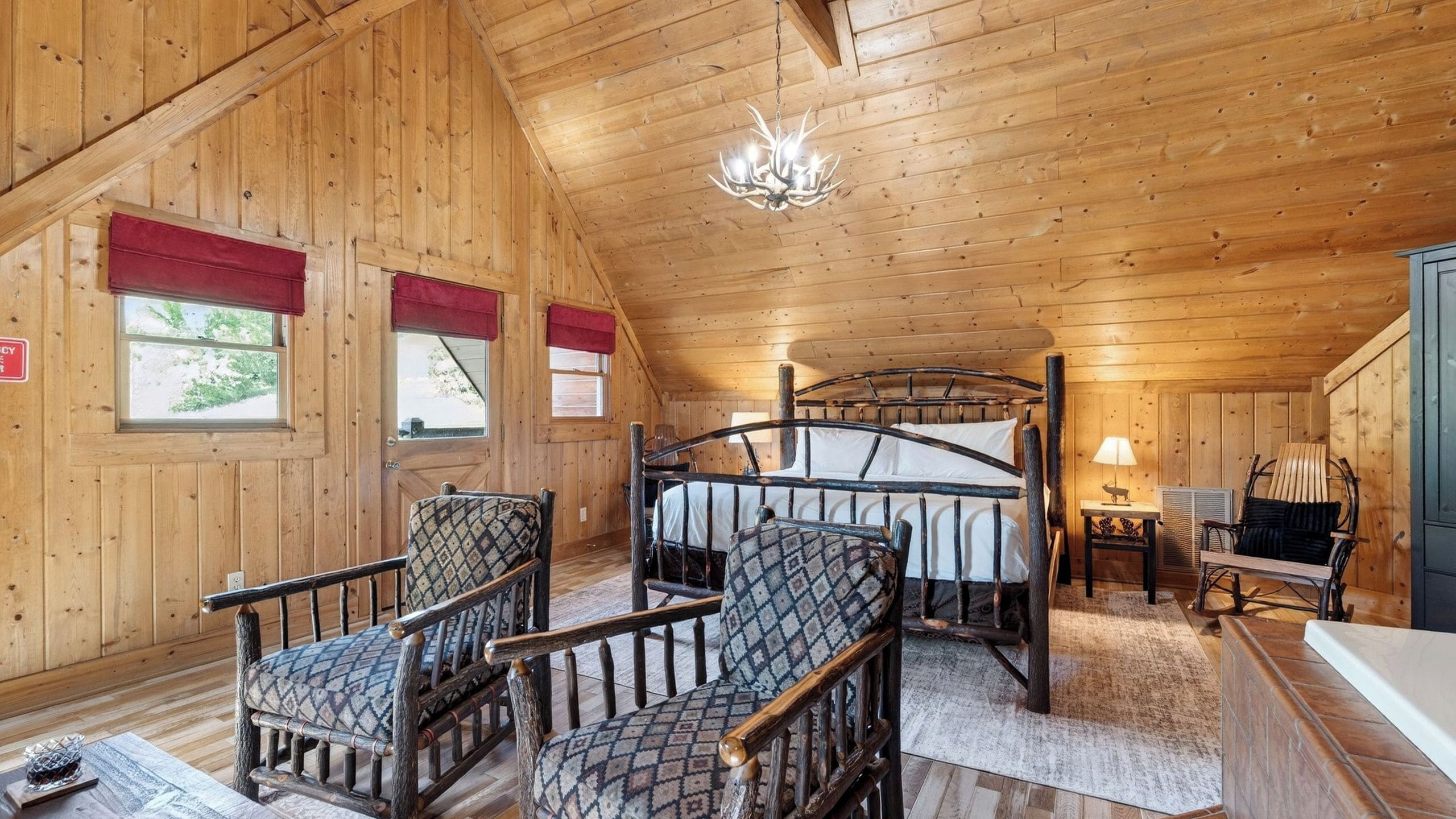 Ein rustikales Blockhaus-Interieur mit einem Bett, zwei gemusterten Sesseln, einer Chandelierleuchtre und einem kleinen Tisch mit einer Lampe.