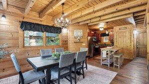 A wooden cabin interior with a dining table, chairs, a chandelier, a kitchen area, and a view through the window. - Smoky Mountain Playhouse by Avantstay Hot Tub + Luxe Cabin + Games + Views Pigeon Forge Area (Sevierville)