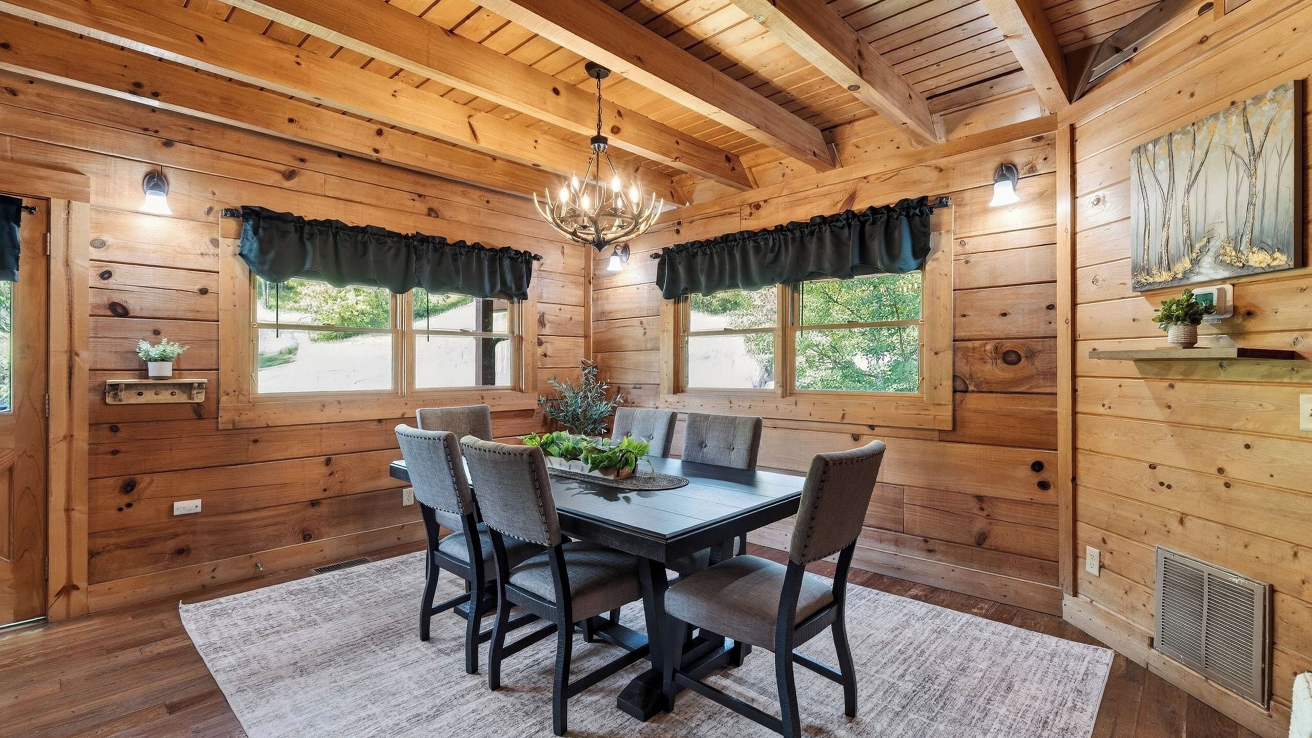 A wooden cabin interior with a dining table, chairs, a chandelier, and windows with curtains.