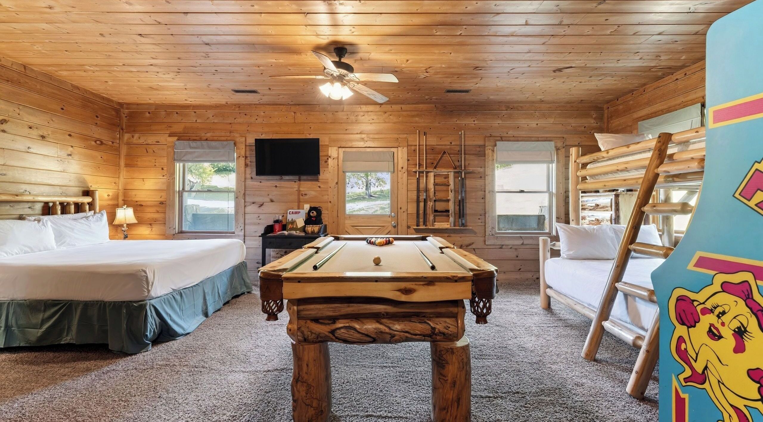 A wooden cabin room with a bed, a pool table, a TV, and a window.