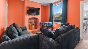A living room with orange walls, a fireplace, a foosball table, and a TV mounted on the wall. - Stylish 3BR Victorian House | Garden, BBQ, Parking (Worcestershire)