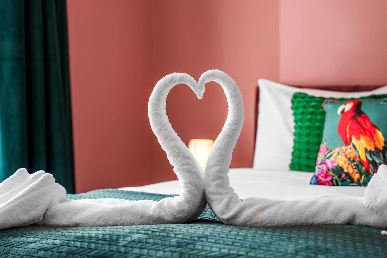 A bed with a heart-shaped towel arrangement, a colorful pillow, and a lamp in the background.