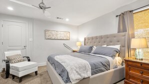 A bedroom with a bed, a chair, a nightstand, a lamp, a ceiling fan, and a wall-mounted artwork.