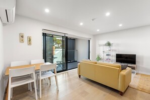 A modern living room with a sofa, a TV, a dining table with chairs, and a sliding glass door leading to an outdoor area. - Modern apartment near the airport (Mangere Bridge)