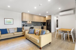A modern living room with a beige sofa, a wooden dining table, and a kitchen area with wooden cabinets and a black countertop. - Modern apartment near the airport (Mangere Bridge)