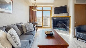 A living room with a sofa, a coffee table, a fireplace, a TV, and a balcony with a view.
