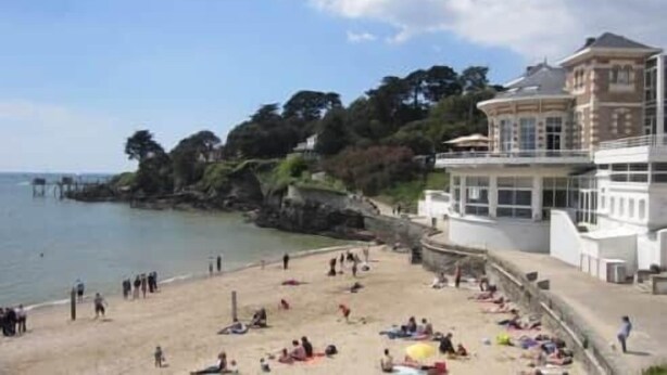 A beach with people sunbathing, a building with a balcony, and a pier in the background. - To complete (Pornic)