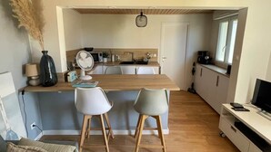 A compact kitchen with a wooden countertop, two white chairs, a dining table, and a TV on a stand. - To complete (Pornic)