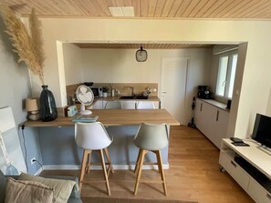 Eine kompakte Küche mit Holzausziehtisch, zwei weißen Stühlen und einem Fernseher auf einem Standfuß.