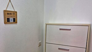 Esquina de una habitación con una pared blanca, un cartel de madera con la inscripción 'WIFI PASSWORD' y un código QR, y un archivador de color claro con tres cajones.