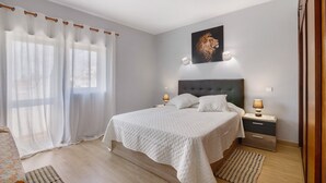 Un dormitori amb un llit, dues taules de nit, una tauleta, una làmpada de paret, una finestra amb cortines i una fotografia amb marc a la paret.