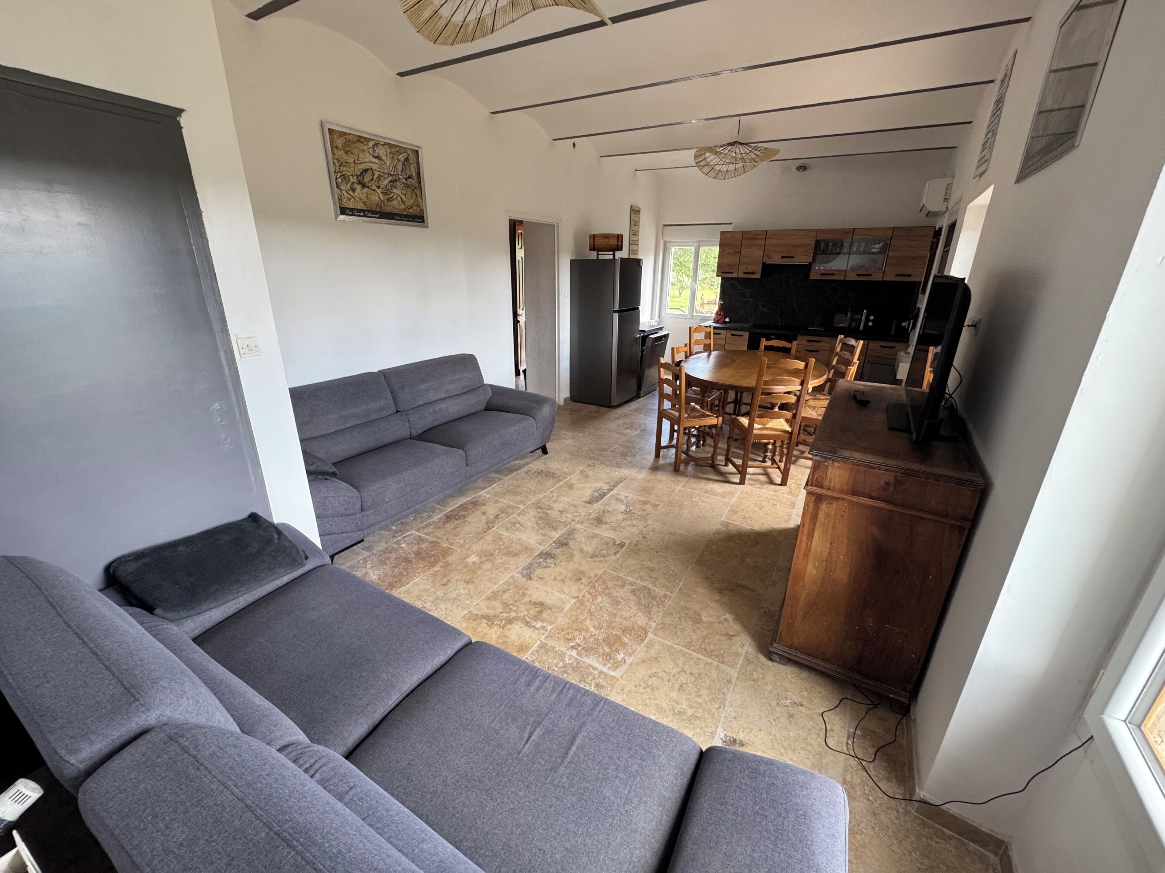 A living room with a grey sofa, a dining area with wooden chairs, a kitchen with wooden cabinets, and a ceiling fan.