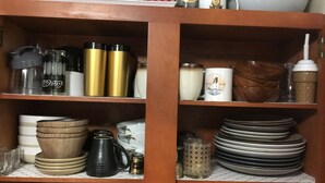 Una repisa de cocina con varios objetos, entre ellos una botella de Coca-Cola, un termo negro y dorado, tazas blancas y platos apilados.